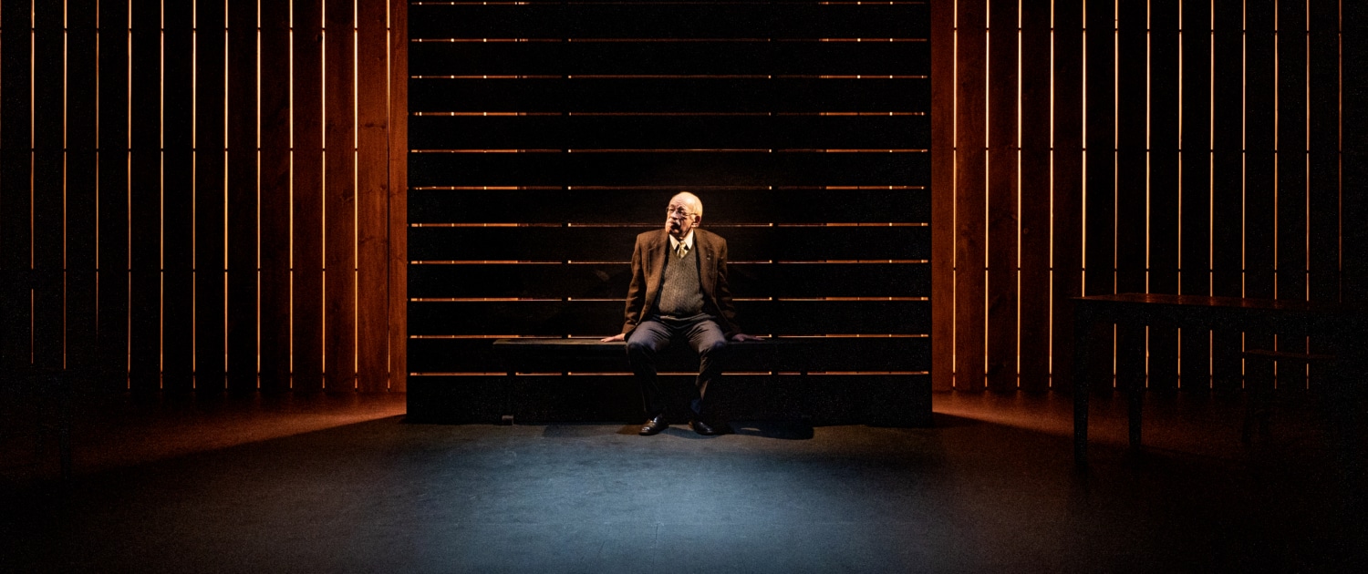 A person sitting on a dark stage with vertical wooden slats in the background, illuminated by a spotlight.