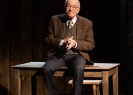 An older man in a jacket, tie and sweater vest sitting on a wooden table with a chair behind, in a dimly lit room with dark wooden walls.