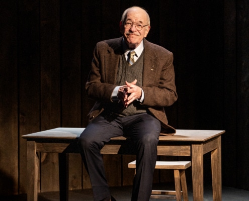 An older man in a jacket, tie and sweater vest sitting on a wooden table with a chair behind, in a dimly lit room with dark wooden walls.