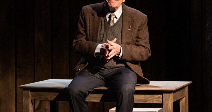 An older man in a jacket, tie and sweater vest sitting on a wooden table with a chair behind, in a dimly lit room with dark wooden walls.