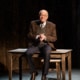 An older man in a jacket, tie and sweater vest sitting on a wooden table with a chair behind, in a dimly lit room with dark wooden walls.