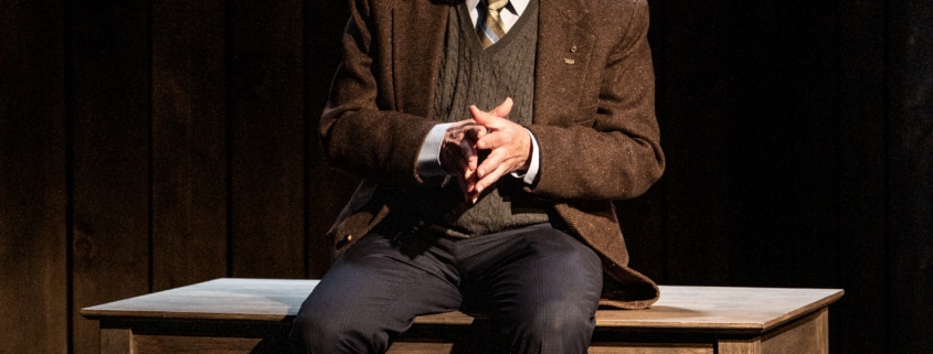 An older man in a jacket, tie and sweater vest sitting on a wooden table with a chair behind, in a dimly lit room with dark wooden walls.