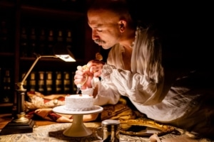 Alberto Denis in a scene from 'Confection' by Third Rail Projects, dressed in historical costume while lighting candles on a small white cake. The setting includes a dimly lit room with bookshelves, a desk lamp, and vintage props like a magnifying glass and ink bottle, evoking a nostalgic and theatrical atmosphere.