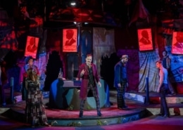 A dramatic theatrical stage scene from a production of 'Assassins,' featuring multiple actors in period costumes. The central figure stands elevated on a platform, holding a staff or cane, surrounded by cast members in dynamic poses. The backdrop includes red-lit posters with silhouetted profiles and text, contributing to a moody, intense atmosphere enhanced by red and blue stage lighting.