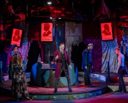 A dramatic theatrical stage scene from a production of 'Assassins,' featuring multiple actors in period costumes. The central figure stands elevated on a platform, holding a staff or cane, surrounded by cast members in dynamic poses. The backdrop includes red-lit posters with silhouetted profiles and text, contributing to a moody, intense atmosphere enhanced by red and blue stage lighting.