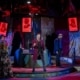 A dramatic theatrical stage scene from a production of 'Assassins,' featuring multiple actors in period costumes. The central figure stands elevated on a platform, holding a staff or cane, surrounded by cast members in dynamic poses. The backdrop includes red-lit posters with silhouetted profiles and text, contributing to a moody, intense atmosphere enhanced by red and blue stage lighting.
