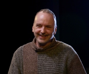 A man wearing a two-toned brown and gray sweater, standing against a dark background.