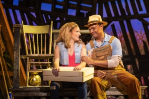 A smiling young woman and older man sit close together on a wooden bench in a warmly lit, rustic barn setting. The woman holds a white box on her lap and wears a patterned denim vest and red top while the man, dressed in overalls and a straw hat, touches her arm affectionately.