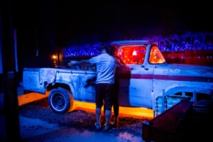 Lia Bonfilio and Ryan Wuestewald in a nighttime scene from 'Sweet & Lucky' by Third Rail Projects, standing beside a rusted vintage pickup truck. The setting is dramatically lit with vibrant blue and orange hues, with a fence and blue-lit foliage in the background, evoking a moody, immersive theatrical atmosphere.