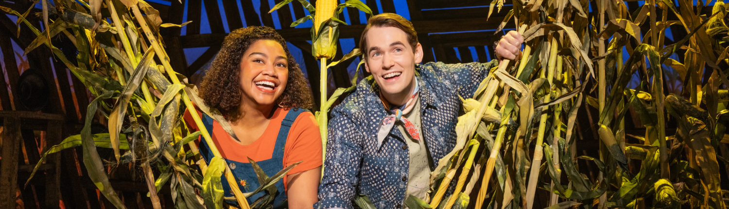Two smiling actors peek through tall corn stalks on a brightly lit stage with a rustin barn backdrop.