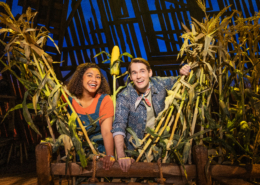 Two smiling actors peek through tall corn stalks on a brightly lit stage with a rustin barn backdrop.