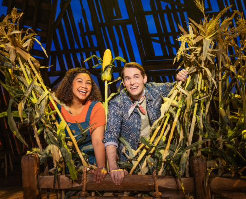 Two smiling actors peek through tall corn stalks on a brightly lit stage with a rustin barn backdrop.