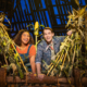 Two smiling actors peek through tall corn stalks on a brightly lit stage with a rustin barn backdrop.