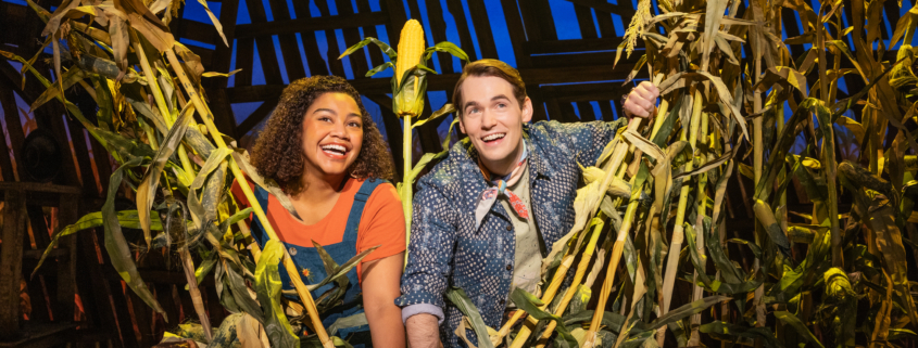 Two smiling actors peek through tall corn stalks on a brightly lit stage with a rustin barn backdrop.