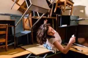 Rachel L. Berman in a scene from 'Then She Fell' by Third Rail Projects, positioned amid a chaotic arrangement of overturned desks and chairs. The furniture is stacked haphazardly, creating a visually intense and unstable environment that reflects the immersive and surreal nature of the performance.