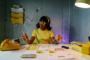 Roxanne Kidd in a scene from 'Behind the City' by Third Rail Projects, seated at a desk in a retro-styled office environment. The setting includes a yellow rotary phone, scattered documents, and classic stationery, with a metal wall in the background featuring file organizers and papers, evoking a mid-century workplace aesthetic.