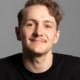 Young man with brown hair and mustache  wearing a black t-shirt and grinning