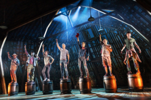 Seven performers stand on barrels with fists raised in a dramatic pose on a barn-themed stage under theatrical lighting.