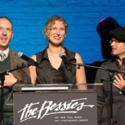 Three individuals standing behind a podium with microphones, presenting at 'The Bessies' - The New York Dance and Performance Awards. The background features a blue brick wall and stage lighting.
