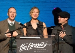 Three individuals standing behind a podium with microphones, presenting at 'The Bessies' - The New York Dance and Performance Awards. The background features a blue brick wall and stage lighting.