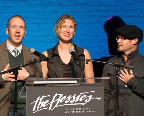 Three individuals standing behind a podium with microphones, presenting at 'The Bessies' - The New York Dance and Performance Awards. The background features a blue brick wall and stage lighting.
