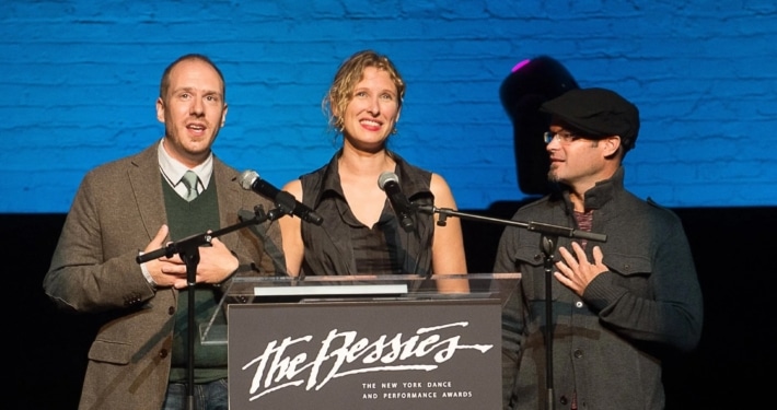 Three individuals standing behind a podium with microphones, presenting at 'The Bessies' - The New York Dance and Performance Awards. The background features a blue brick wall and stage lighting.