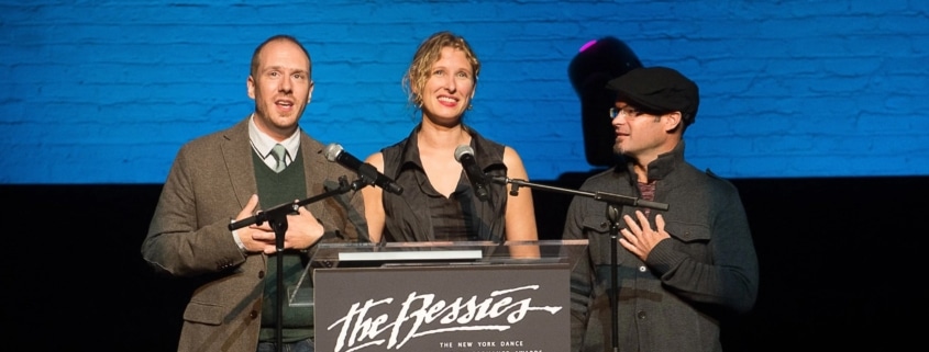Three individuals standing behind a podium with microphones, presenting at 'The Bessies' - The New York Dance and Performance Awards. The background features a blue brick wall and stage lighting.