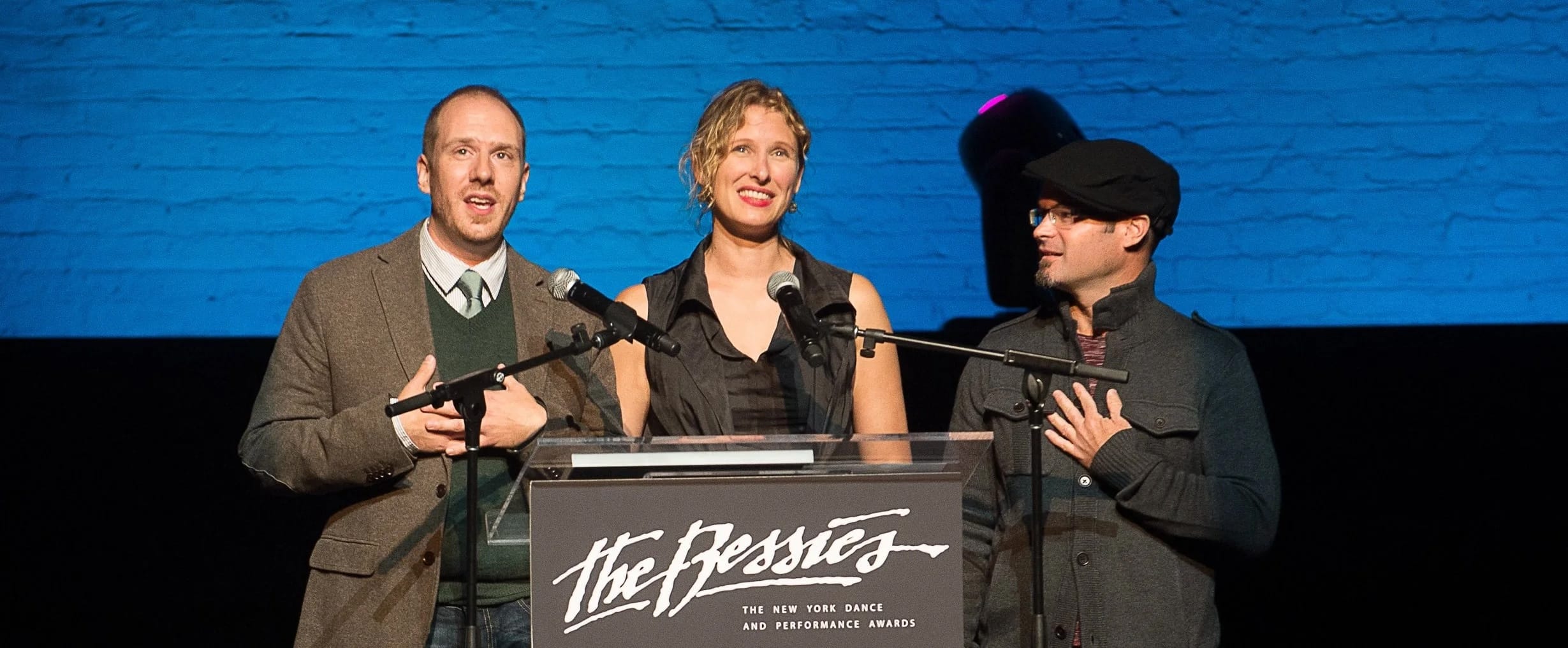 Three individuals standing behind a podium with microphones, presenting at 'The Bessies' - The New York Dance and Performance Awards. The background features a blue brick wall and stage lighting.