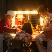 Three performers backstage in. costume applying makeup in front of brightly lit vanity.