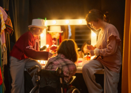 Three performers backstage in. costume applying makeup in front of brightly lit vanity.