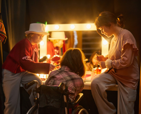 Three performers backstage in. costume applying makeup in front of brightly lit vanity.