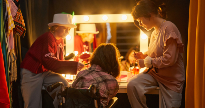 Three performers backstage in. costume applying makeup in front of brightly lit vanity.
