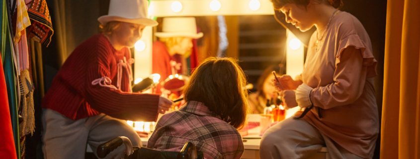 Three performers backstage in. costume applying makeup in front of brightly lit vanity.