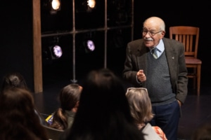 Actor Kenneth Tigar, playing Holocaust survivor Eddie Jaku, stands on a stage as he speaks to an audience.