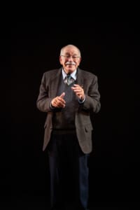 Actor Kenneth Tigar, playing Holocaust survivor Eddie Jaku, stands alone on stage with a dark background, using expressive hand movements to connect with an unseen audience.
