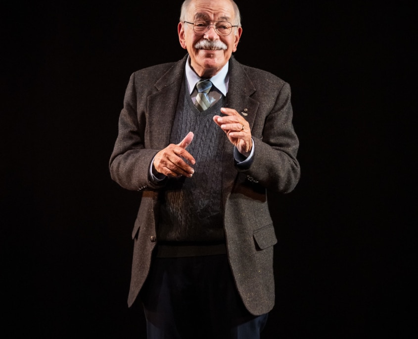 Actor Kenneth Tigar, playing Holocaust survivor Eddie Jaku, stands alone on stage with a dark background, using expressive hand movements to connect with an unseen audience.
