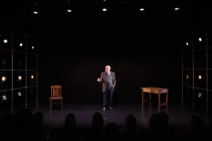 Actor Kenneth Tigar, playing Holocaust survivor Eddie Jaku, stands on stage, addressing the audience. The stage is bare apart from a wooden table and chair.