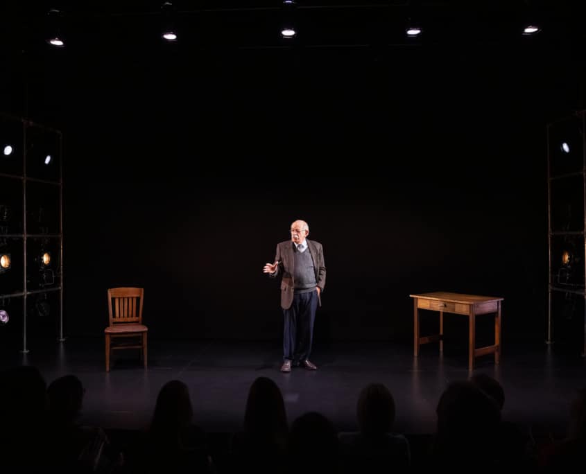 Actor Kenneth Tigar, playing Holocaust survivor Eddie Jaku, stands on stage, addressing the audience. The stage is bare apart from a wooden table and chair.