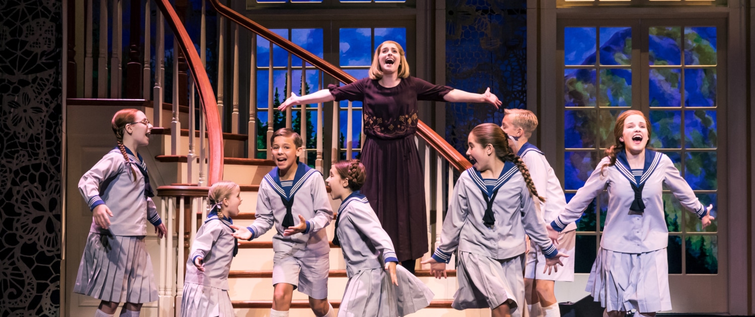  group of children in sailor-style uniforms and a woman in a dark dress are singing and dancing on stage in front of a staircase with large windows showing a scenic backdrop.
