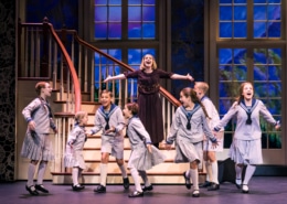 group of children in sailor-style uniforms and a woman in a dark dress are singing and dancing on stage in front of a staircase with large windows showing a scenic backdrop.