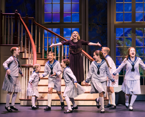 group of children in sailor-style uniforms and a woman in a dark dress are singing and dancing on stage in front of a staircase with large windows showing a scenic backdrop.