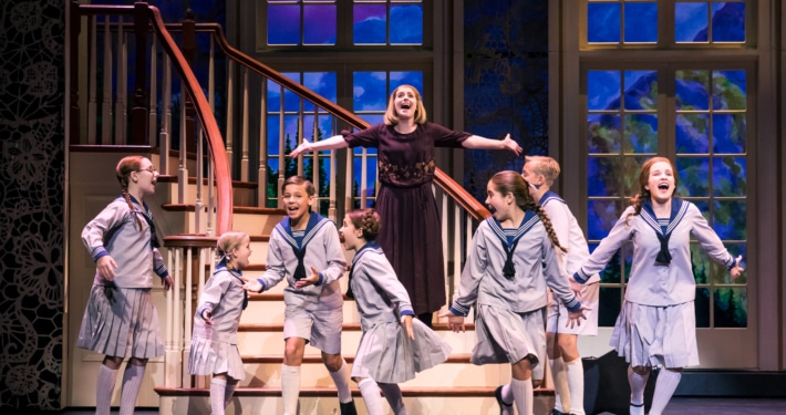group of children in sailor-style uniforms and a woman in a dark dress are singing and dancing on stage in front of a staircase with large windows showing a scenic backdrop.