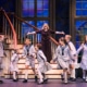 group of children in sailor-style uniforms and a woman in a dark dress are singing and dancing on stage in front of a staircase with large windows showing a scenic backdrop.