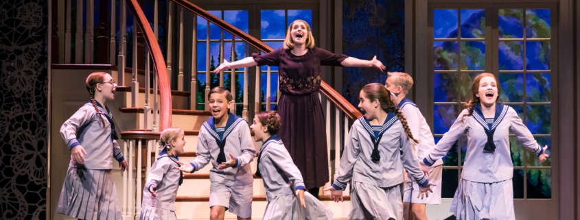 group of children in sailor-style uniforms and a woman in a dark dress are singing and dancing on stage in front of a staircase with large windows showing a scenic backdrop.