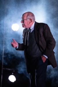 Actor Kenneth Tigar, playing Holocaust survivor Eddie Jaku, is mid-speech, framed by atmospheric smoke effects and backlighting, creating a visually striking and dramatic scene.