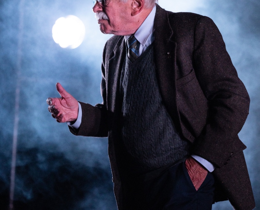 Actor Kenneth Tigar, playing Holocaust survivor Eddie Jaku, is mid-speech, framed by atmospheric smoke effects and backlighting, creating a visually striking and dramatic scene.