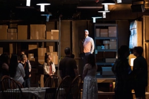 A warehouse with boxes and dim lighting. In it are several people looking toward a man standing atop a table in a moment from Sweet and Lucky: Echo