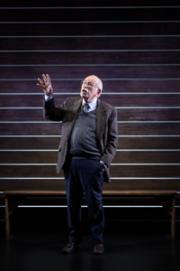 Actor Kenneth Tigar, playing Holocaust survivor Eddie Jaku, stands against a wooden backdrop with one hand raised.