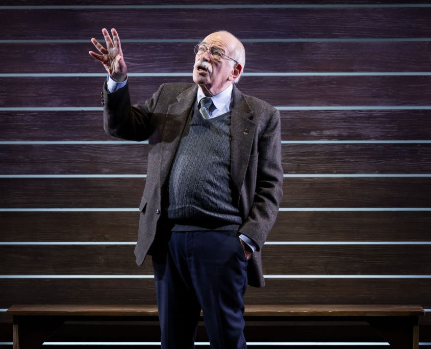 Actor Kenneth Tigar, playing Holocaust survivor Eddie Jaku, stands against a wooden backdrop with one hand raised.