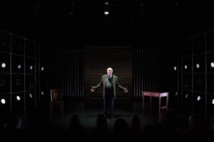 Actor Kenneth Tigar, playing Holocaust survivor Eddie Jaku, stands on a stage under dramatic lighting with shadows cast around them, addressing an unseen crowd.